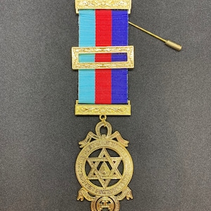Puede incluir: Pendiente masónico de oro con una estrella de seis puntas y un círculo de texto alrededor. El pendiente está unido a una cinta azul, roja y blanca con una barra dorada.