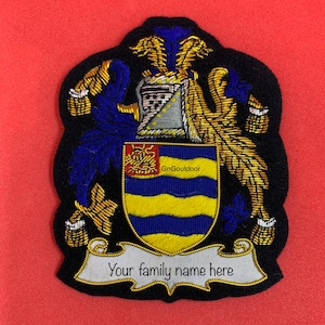 Könnte beinhalten: Ein schwarzer gestickter Wappen mit goldenen und blauen Details. Das Wappen zeigt einen Schild mit einem gelben und blauen Streifenmuster, eine Krone und eine Schriftrolle mit dem Text "Ihr Familienname hier".