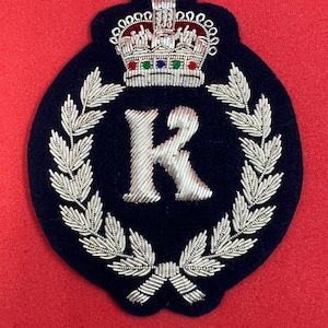 May include: A navy blue embroidered patch with a silver K surrounded by a silver laurel wreath and a silver crown with red, green, and blue jewels.