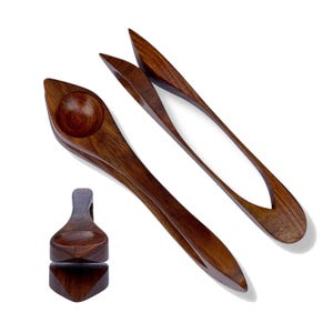 May include: A selection of handcrafted wooden kitchen utensils. The set includes a spoon with a rounded bowl, a pair of tongs, and a small, geometric-shaped item. The wood has a rich, dark brown colour and a smooth finish.