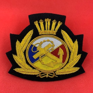 Könnte beinhalten: Ein schwarzer gestickter Aufnäher mit einem goldenen Anker, einem Seil und einem Lorbeerkranz. Der Anker ist von einem Kreis mit einem rot-weiß-blauen Design umgeben. Eine goldene Krone sitzt über dem Anker.