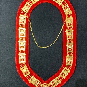 Può includere: Una catena d'oro con un retro in velluto rosso. La catena è composta da maglie ripetute con un motivo a stella e mezzaluna. La catena ha la forma di un collare.