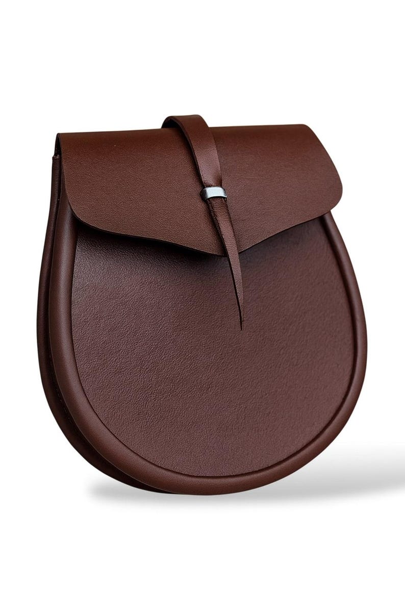 May include: A brown leather bag with a curved, rounded bottom and a flap closure. The bag has a strap with a silver-colored metal accent. The bag is made of a textured leather material.