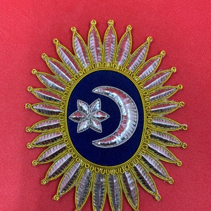 May include: A silver and gold embroidered patch with a crescent moon and star on a blue background. The patch is surrounded by a circle of gold and silver leaves.