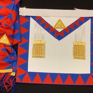 Könnte beinhalten: Eine weiße Freimaurer-Schürze mit rotem und blauem Rand. Die Schürze hat zwei weiße Bänder mit goldenen Quasten. Die Schürze hat ein goldenes Dreieck mit dem Buchstaben 'G' in der Mitte.