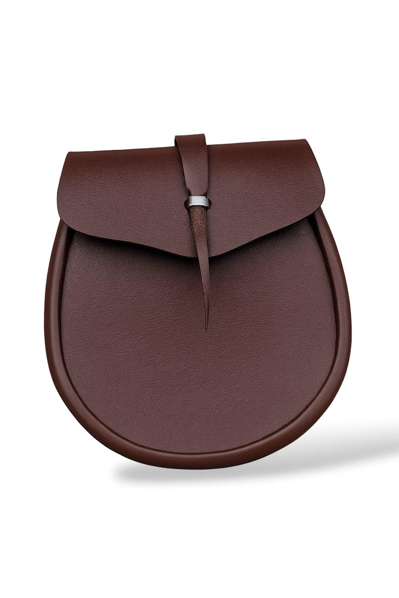 May include: A brown leather sporran with a rounded bottom and a flap closure. The flap is secured with a leather strap and a silver-colored metal ring. The sporran has a textured surface.