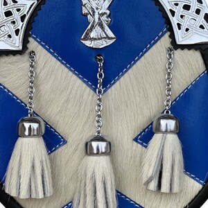 May include: A blue and white kilt sporran with three white tassels and a silver metal embellishment featuring a cross and a figure.
