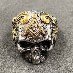 Puede incluir: Un anillo de calavera de plata con un símbolo masónico dorado en la frente. La calavera está detallada con una superficie texturizada y el símbolo está rodeado de alas doradas.