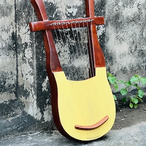 Pode incluir: Uma lira de madeira com caixa de ressonância amarela e detalhes castanhos. O instrumento tem cordas e uma ponte de madeira curva. A lira está encostada numa parede cinzenta texturizada.