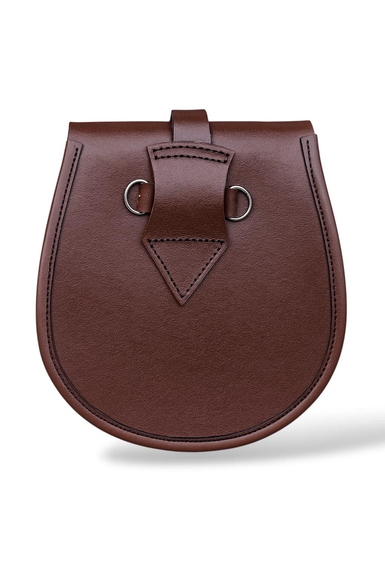 May include: A brown leather sporran with a rounded bottom and a flap closure. The sporran has a decorative stitched border and silver-colored metal rings. The flap is secured with a leather strap.