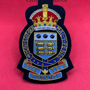 May include: Embroidered patch featuring a royal crown and intricate detailing in gold, red, blue, and black. The patch includes text in gold lettering and is set against a red background. The patch is likely a military or organizational emblem.
