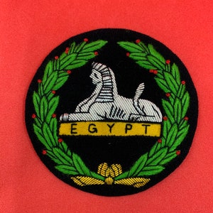 Puede incluir: Un parche circular bordado negro con una esfinge plateada, una pancarta dorada con la palabra "EGIPTO" y una corona de laurel verde.