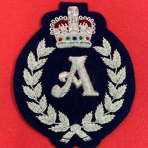 Puede incluir: Un parche bordado azul marino con la letra "A" plateada dentro de una corona de laurel. Una corona plateada con detalles rojos, verdes y azules se encuentra encima de la letra. El parche está sobre un fondo rojo.