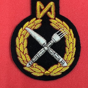 May include: A black embroidered patch with a gold wreath surrounding a silver knife and fork crossed over each other. The wreath has a red outline.