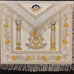 May include: White Masonic apron with gold embroidery. The apron features two pillars, a compass and square, and an all-seeing eye. The apron is trimmed with silver fringe.