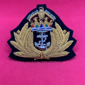 Op de afbeelding: Een gouden geborduurde patch met een zilveren anker, een kroon en een krans van bladeren op een zwarte achtergrond.