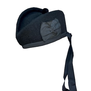 Op de afbeelding: Een zwarte baret met een strik en lintdetail. De hoed heeft een afgeronde bovenkant en een kleine pompon. Het lint hangt van de zijkant van de hoed af. Een klassieke hoed.