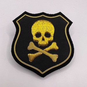Puede incluir: Un parche bordado negro en forma de escudo con un diseño de calavera y huesos cruzados dorados.