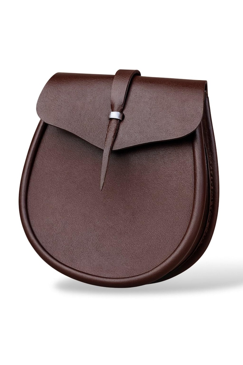 May include: A brown leather sporran with a curved bottom and a flap closure. The flap is secured with a leather strap and a silver-colored metal ring. The sporran is a traditional Scottish accessory.