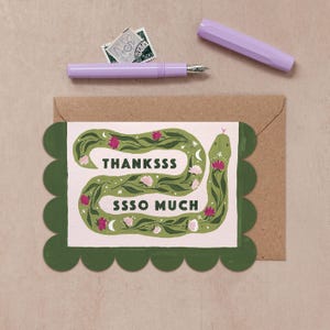 May include: A rectangular greeting card with a scalloped dark green border features a green snake illustration on a light pink background. The text reads THANKSSS SSSO MUCH. A light purple fountain pen and a postage stamp are placed above the card.