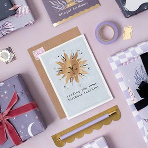 The gently smiling sun bursts from the birthday card in glittering gold foil, peeking at the birthday card caption which reads Sending you some birthday sunshine in a hand lettered font. The birthday card has light blue background and kraft envelope.