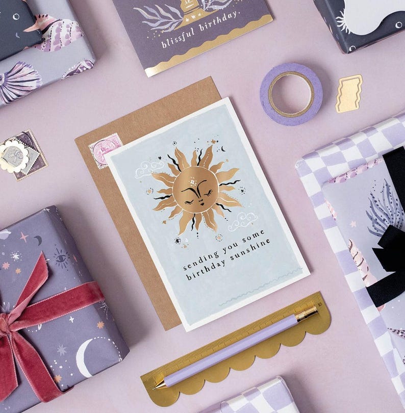 The gently smiling sun bursts from the birthday card in glittering gold foil, peeking at the birthday card caption which reads Sending you some birthday sunshine in a hand lettered font. The birthday card has light blue background and kraft envelope.
