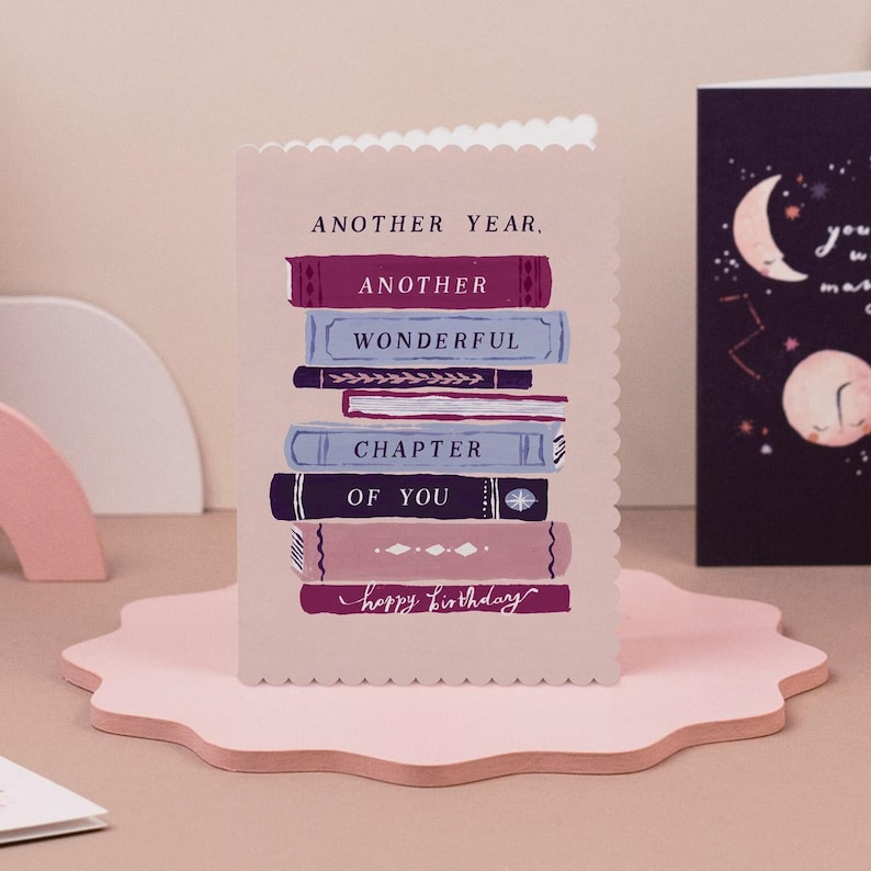 A birthday card with a stack of books and the text "Another Year, Another Wonderful Chapter of You" printed on it, placed on a pink surface.