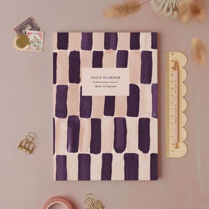 Könnte beinhalten: Ein Daily Planner mit einem geometrischen Muster aus lila und beigen Pinselstrichen. Das Cover trägt den Text "DAILY PLANNER" und "Made in England". Ein rosa Lineal und Bleistift befinden sich auf der rechten Seite.