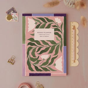 Könnte beinhalten: Ein Wochenplaner mit floralem Design in Rosa, Grün und Weiß. Auf dem Cover steht "WEEKLY PLANNER" und "Made in England". Ein Lineal und ein Bleistift befinden sich auf der rechten Seite des Planers. Dekorative Elemente sind Briefmarken und eine goldene Büroklammer.