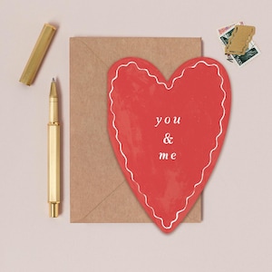 a heart-shaped card with the words "you & me" written on it, placed on a brown envelope, along with a pen and some postage stamps.