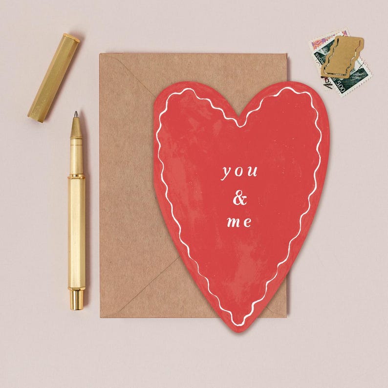 a heart-shaped card with the words "you & me" written on it, placed on a brown envelope, along with a pen and some postage stamps.