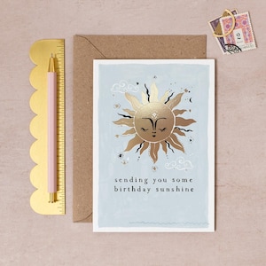 The gently smiling sun bursts from the birthday card in glittering gold foil, peeking at the birthday card caption which reads Sending you some birthday sunshine in a hand lettered font. The birthday card has light blue background and kraft envelope.