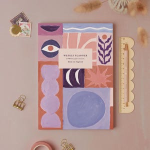 Peut inclure: Un agenda hebdomadaire avec un design abstrait dans les tons de violet, rose et bleu. La couverture présente des formes géométriques, un œil et le texte "WEEKLY PLANNER Made in England". Une règle et un crayon roses sont à droite.