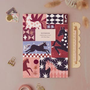 May include: A rectangular notebook with a multi-paneled cover featuring illustrations of horses in shades of pink, red, purple, and black, with stars and geometric patterns. The text reads NOTEBOOK and 192 Plain Pages. A 15 cm ruler is visible.