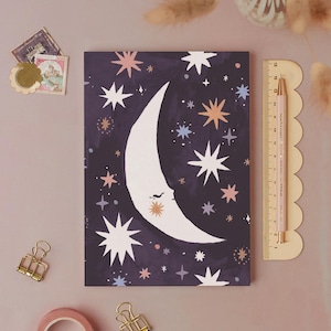 May include: A rectangular notebook with a dark purple cover featuring a white crescent moon with a closed eye and various white, pink, and light blue stars. A wooden ruler measuring 15 cm is placed vertically next to the notebook.