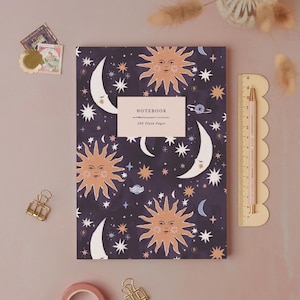 Könnte beinhalten: Ein Notizbuch mit Himmelsmotiven und einem dunkelblauen Hintergrund, das Illustrationen von Sonne, Mond und Sternen zeigt. Das Notizbuch hat ein weißes Etikett mit der Aufschrift "NOTEBOOK 192 Plain Pages". Ein rosa Bleistift und Lineal befinden sich auf der rechten Seite.
