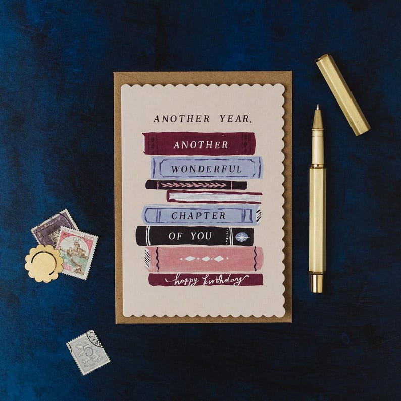 A birthday card with a stack of books and a pen, along with some stamps and a gold pen, all placed on a blue background.