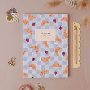 Könnte beinhalten: Ein rechteckiges Notizbuch mit hellblau-weißem Karomuster und Illustrationen von Croissants und Erdbeeren. Ein zentrales Etikett trägt die Aufschrift NOTEBOOK und 192 Plain Pages. Ein Lineal zeigt eine Länge von etwa 20 cm.
