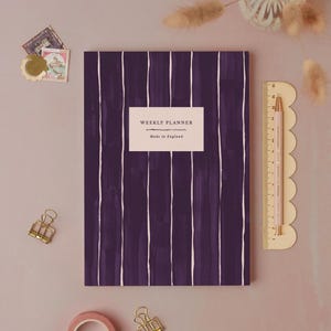 Peut inclure: Un agenda hebdomadaire violet avec des coups de pinceau verticaux blancs. L'agenda porte une étiquette rectangulaire indiquant « WEEKLY PLANNER Made in England ». Une règle et un crayon roses sont à droite. Des trombones dorés et des timbres décoratifs sont également présents.