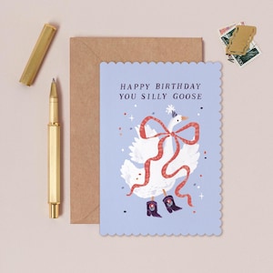 May include: A light blue birthday card with a scalloped edge features a whimsical illustration of a goose wearing a party hat and boots, with a red bow. The text reads "HAPPY BIRTHDAY YOU SILLY GOOSE". A gold pen and envelope are next to the card.