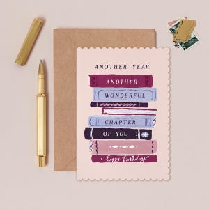 Puede incluir: Una tarjeta de cumpleaños con una pila de libros ilustrados en tonos morados, azules y rosas, con las palabras "Another Year, Another Wonderful Chapter of You" y "Happy Birthday". Un bolígrafo y un sobre dorados están junto a la tarjeta.