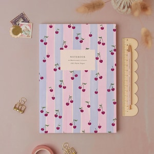 Può includere: Un quaderno con un motivo a righe rosa e blu e illustrazioni di ciliegie. La copertina presenta la scritta "NOTEBOOK" e il testo "192 Plain Pages". Una matita rosa e un righello sono accanto al quaderno.