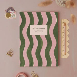 May include: A weekly planner with a green and pink wavy pattern. The planner has the text "WEEKLY PLANNER" and "Made in England" on a white label. A pink ruler and pencil are on the right. Decorative items like stamps and paper clips are also present.