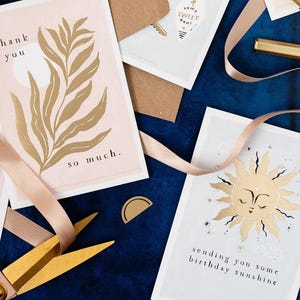 The gently smiling sun bursts from the birthday card in glittering gold foil, peeking at the birthday card caption which reads Sending you some birthday sunshine in a hand lettered font. The birthday card has light blue background and kraft envelope.