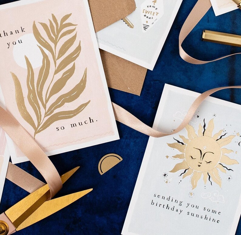 The gently smiling sun bursts from the birthday card in glittering gold foil, peeking at the birthday card caption which reads Sending you some birthday sunshine in a hand lettered font. The birthday card has light blue background and kraft envelope.