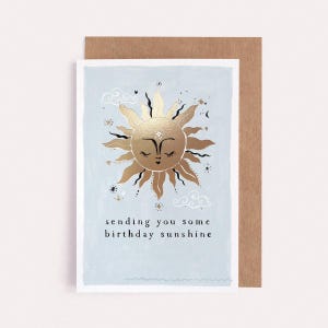 The gently smiling sun bursts from the birthday card in glittering gold foil, peeking at the birthday card caption which reads Sending you some birthday sunshine in a hand lettered font. The birthday card has light blue background and kraft envelope.