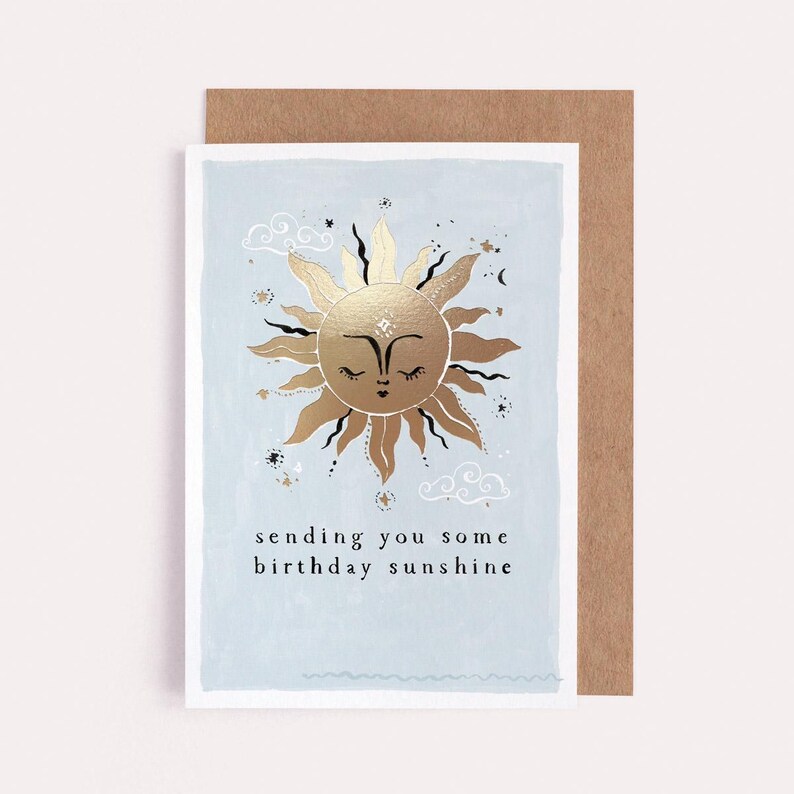 The gently smiling sun bursts from the birthday card in glittering gold foil, peeking at the birthday card caption which reads Sending you some birthday sunshine in a hand lettered font. The birthday card has light blue background and kraft envelope.