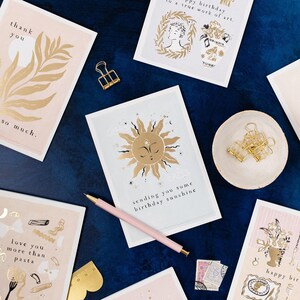 The gently smiling sun bursts from the birthday card in glittering gold foil, peeking at the birthday card caption which reads Sending you some birthday sunshine in a hand lettered font. The birthday card has light blue background and kraft envelope.