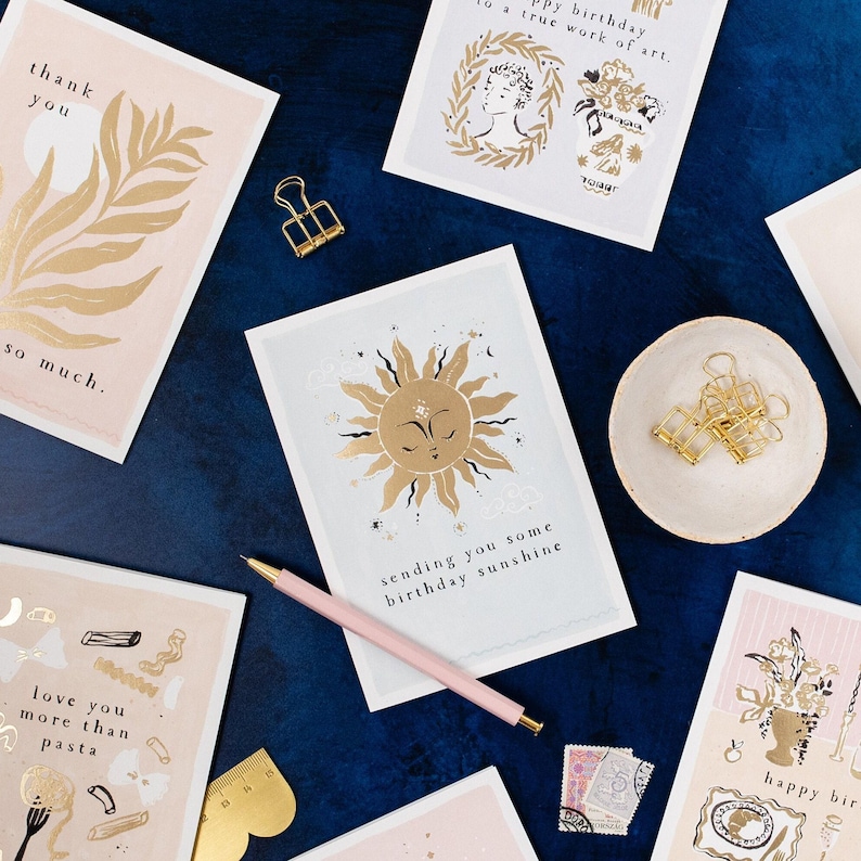 The gently smiling sun bursts from the birthday card in glittering gold foil, peeking at the birthday card caption which reads Sending you some birthday sunshine in a hand lettered font. The birthday card has light blue background and kraft envelope.