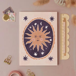 May include: A rectangular notebook with a light pink cover featuring a central dark purple oval containing a tan sun face with rays, surrounded by white stars and a crescent moon. A 15 cm ruler, two gold binder clips, and two postage stamps are present.